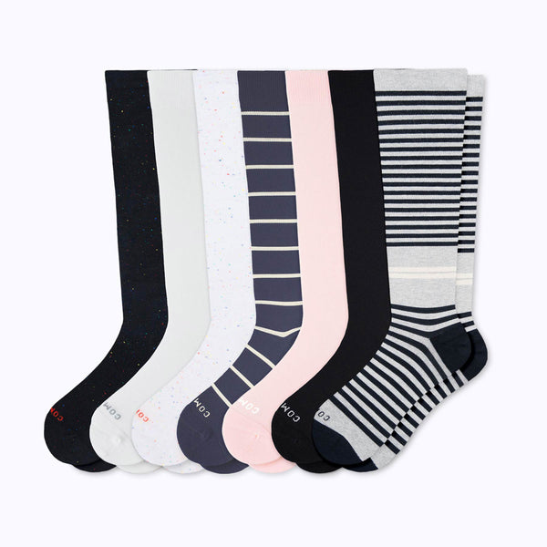 Seven pairs of knee-high socks arranged in a fan shape on a light gray background. The socks feature various patterns including solid black, white speckles, navy blue stripes, light pink stripes, and black and white horizontal stripes.