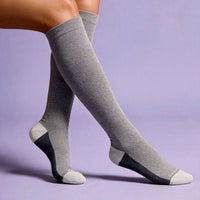 A pair of gray knee-high compression socks with black and white accents on the heels, toes, and heels, shown on a model's legs against a solid lavender background.