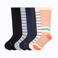 7 pairs of knee-high socks on a light grey background: 1 black speckled, 1 solid black, 1 grey with charcoal stripes, 1 navy solid, 1 charcoal grey solid, 1 rose with navy stripes, 1 cream with terra cotta stripes.