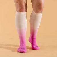 A pair of knee-high compression socks with a berry to white ombre design on a model standing on a beige background