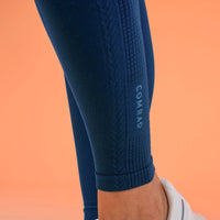Close-up of a person's lower leg wearing dark blue textured compression leggings with a vertical COMRAD logo on the calf, paired with a white sneaker against a solid orange background.