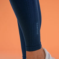 Close-up of a person's lower leg wearing dark navy textured navy compression leggings with a vertical COMRAD logo on the calf, paired with a white shoe against a solid orange background.
