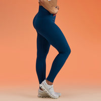 A model wearing high-waisted, form-fitting compression leggings in a deep navy blue, paired with white sneakers, posed against a solid peach-orange studio background.