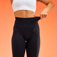 A close-up of a model wearing black high-waisted leggings, pulling the waistband outward to demonstrate stretch. The model is also wearing a white sports bra, set against a solid orange background.