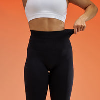 A model wearing black high-waisted leggings and a white sports bra, pulling the waistband of the leggings to demonstrate stretch, against a solid orange background.