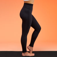 A side-profile view of a person wearing black high-waisted ribbed leggings, standing barefoot on a black mat against a solid orange background.