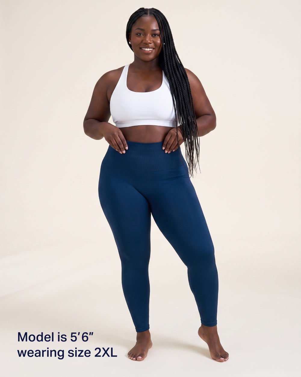 A woman modeling high-waisted navy blue leggings with a white sports bra, standing against a light beige studio background. Text in the bottom left corner reads 'Model is 5'6" wearing size 2XL'.