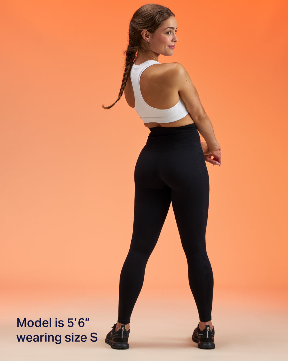 A woman modeling black high-waisted leggings and a white sports bra against a solid orange background, shown from the back with text in the corner stating 'Model is 5'6" wearing size S'.