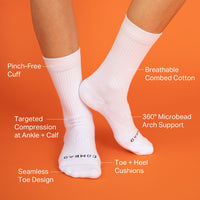 A pair of white crew socks worn on feet against a solid orange background, with multiple text callouts highlighting product features like pinch-free cuff, breathable combed cotton, targeted compression, and toe cushioning.